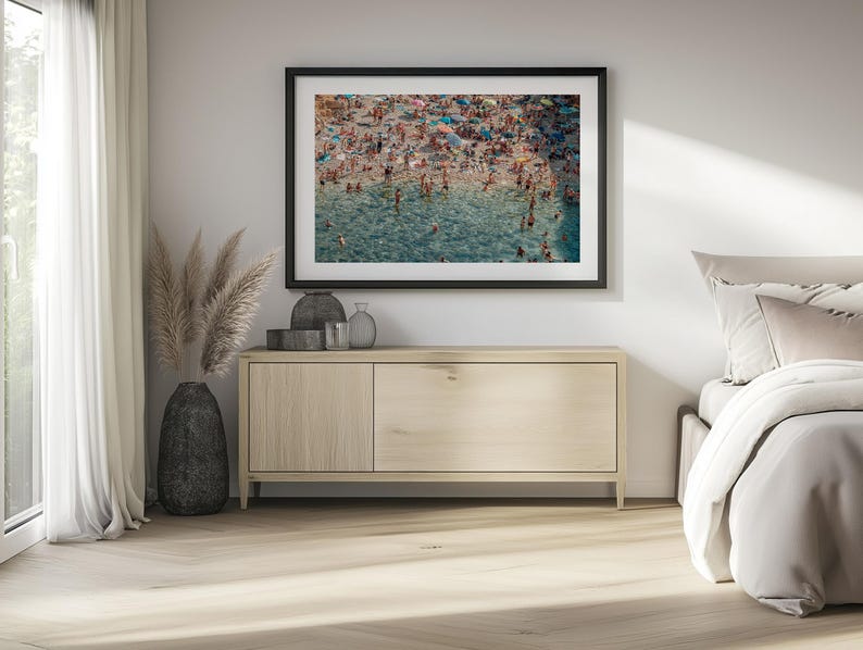 May include: A framed photograph of a crowded beach scene with people swimming and sunbathing in the turquoise water. The photo is hanging on a white wall above a light wood dresser.