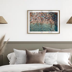 May include: A framed photograph of a crowded beach with people swimming and sunbathing in the turquoise water. The beach is surrounded by a sandy shore and lush green vegetation.