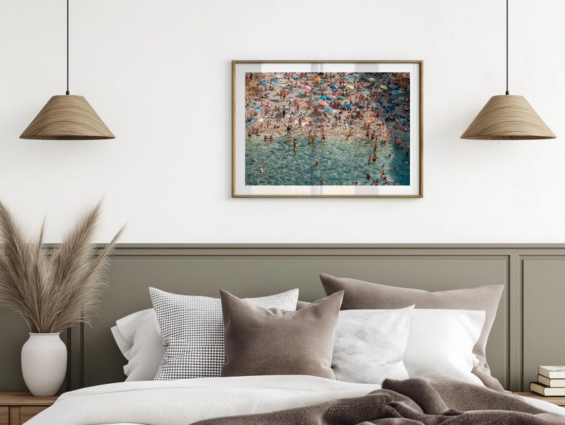 May include: A framed photograph of a crowded beach with people swimming and sunbathing in the turquoise water. The beach is surrounded by a sandy shore and lush green vegetation.