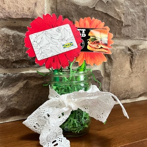 Puede incluir: Dos tarjetas de regalo, una para IKEA y otra para una tienda de s&aacute;ndwiches, est&aacute;n unidas a flores de papel rojas y naranjas en un frasco de vidrio con papel triturado verde y encaje blanco.