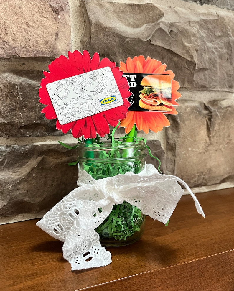 Puede incluir: Dos tarjetas de regalo, una para IKEA y otra para una tienda de s&aacute;ndwiches, est&aacute;n unidas a flores de papel rojas y naranjas en un frasco de vidrio con papel triturado verde y encaje blanco.
