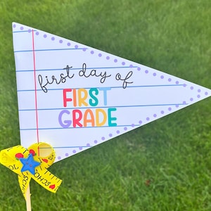 Op de afbeelding: Een witte vlag met een blauwe, groene, gele en paarse "First Grade" banner en de tekst "first day of" in zwart. De vlag is versierd met een paarse gestippelde rand en is bevestigd aan een gele meetlint met een blauwe ster en een rode appel.