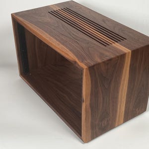 May include: A rectangular wooden cabinet with a dark brown finish and a lighter brown accent. The top features a series of parallel slots. The cabinet has an open front and a black metal strip on the inside.
