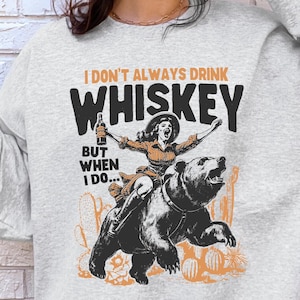 May include: A gray sweatshirt with the text 'I don't always drink whiskey but when I do...' and a cartoon image of a woman riding a bear. The woman is wearing a cowboy hat and holding a bottle of whiskey.