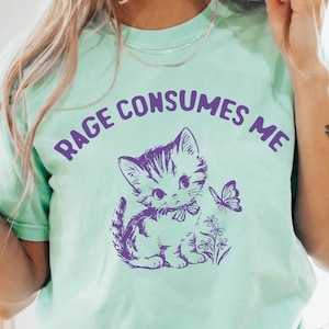 Puede incluir: Una camiseta verde claro con el texto "Rage Consumes Me" en letras moradas. Un gatito de dibujos animados con una mariposa est&aacute; impreso en la camiseta.