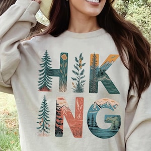 Puede incluir: Sudadera de color arena con la palabra "HIKING" en letras coloridas con tem&aacute;tica de la naturaleza. Las letras est&aacute;n dise&ntilde;adas con im&aacute;genes de &aacute;rboles, monta&ntilde;as y paisajes. La sudadera es de color neutro y tiene mangas largas.