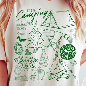 Może przedstawiać: Kremowa koszulka z zieloną grafiką o tematyce kempingowej. Projekt zawiera tekst "Let's Go Camping" i "Adventure Awaits", a także ilustracje namiotu, ogniska i innych niezbędnych element&oacute;w kempingowych.