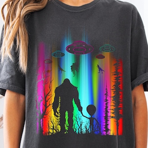 Puede incluir: Una camiseta negra con un estampado gráfico de un Bigfoot y un pequeño extraterrestre de pie en un bosque con un fondo de arcoíris y varios ovnis en el cielo.