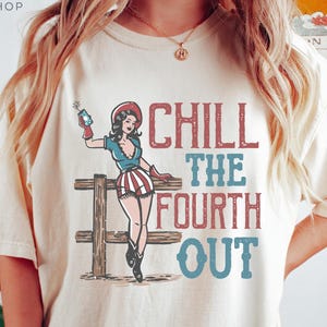 May include: Ivory-colored t-shirt with a graphic of a woman in patriotic attire holding a firecracker, leaning on a wooden fence. The text "CHILL THE FOURTH OUT" is printed in red and blue.