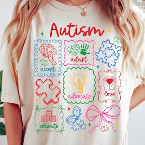 Puede incluir: Camiseta de color marfil con la palabra "Autismo" en escritura roja. La camiseta presenta cuadrados gráficos coloridos con palabras como "aceptar", "adaptar" y "amar", junto con piezas de rompecabezas y una ilustración de cerebro.