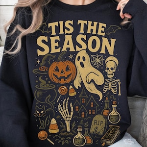 May include: Black sweatshirt featuring a Halloween design with a pumpkin, ghost, and skeleton. The text "TIS THE SEASON" is in gold. Other elements include a witch's hat, candy corn, and a haunted house. The sweatshirt is black.