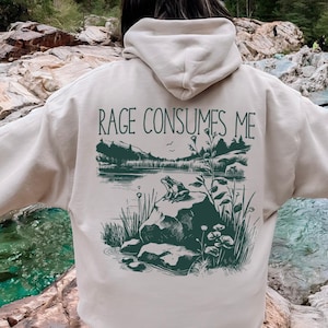 May include: Sand-colored hoodie with the text "RAGE CONSUMES ME" above a green illustration of a frog on a rock by a lake. The hoodie has a hood and long sleeves. The word "SAND" is printed at the bottom.