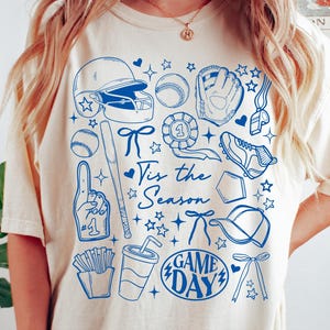 以下が含まれることがあります： アイボリー色のTシャツに、青い野球をテーマにしたグラフィック。デザインには、ヘルメット、野球ボール、グローブ、バット、「Tis the Season」と「Game Day」の文字が含まれています。カジュアルな半袖スタイルです。