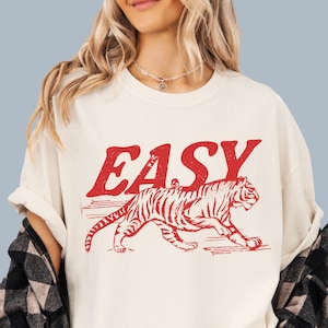 Może przedstawiać: Kremowa koszulka z napisem "EASY" w czerwonych literach nad czerwoną ilustracją tygrysa. Tygrys jest przedstawiony w ruchu, z widocznymi paskami. Koszulka ma luźny kr&oacute;j.