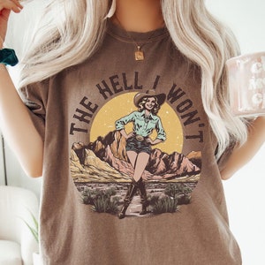May include: Brown t-shirt with a graphic of a woman in a cowboy hat and jeans standing in a desert landscape. The text "The Hell I Won't" is written around the graphic.