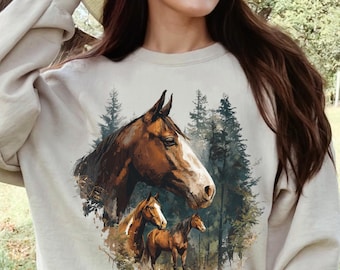 May include: Sand-colored sweatshirt featuring a detailed illustration of horses in a forest setting. The design includes a large horse head and two smaller horses, with a backdrop of evergreen trees. The word "SAND" is printed at the bottom.