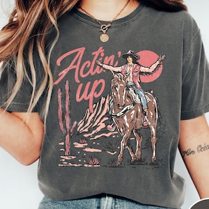 May include: Dark grey t-shirt with a graphic of a person riding a horse in a desert scene. The text "Actin' Up" is in pink script. The person is wearing a cowboy hat and vest. A cactus and sun are in the background.