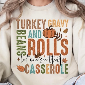 May include: Sand-colored sweatshirt with the text "TURKEY GRAVY AND ROLLS" and "Let me see that CASSEROLE" in various colors. A pumpkin and fall leaves are also featured in the design.