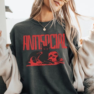 Może przedstawiać: Szary t-shirt z czerwonym nadrukiem graficznym przedstawiającym czarnego kota siedzącego obok czaszki. Tekst "ANTISOCIAL" jest nadrukowany na czerwono nad kotem i czaszką.