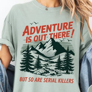 以下が含まれることがあります： 「ADVENTURE IS OUT THERE！」の赤い文字がプリントされた、山と森のグラフィックが特徴のライトグリーンのTシャツ。グラフィックの下には赤い文字で「BUT SO ARE SERIAL KILLERS」と書かれています。半袖のシャツです。