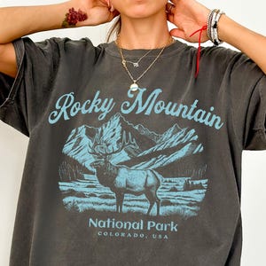 Puede incluir: Camiseta gris oscuro con un gráfico azul de montañas, un alce y el texto "Rocky Mountain National Park Colorado, USA". La camiseta tiene un aspecto vintage y desgastado.