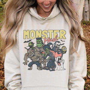 Puede incluir: Una sudadera con capucha beige claro con un estampado gr&aacute;fico de monstruos de dibujos animados. El texto "Monster Mash" est&aacute; impreso en amarillo y rojo en la sudadera.