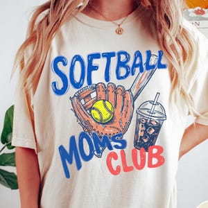May include: Ivory-colored t-shirt with the words "SOFTBALL MOMS CLUB" in blue and red. The design includes a softball, glove, and a drink with ice. The shirt is a casual style.