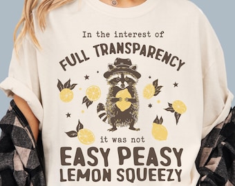 May include: A cream colored t-shirt with a raccoon graphic holding a lemon. The text on the shirt reads "In the interest of full transparency it was not easy peasy lemon squeezy".