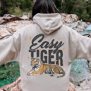 Puede incluir: Una sudadera con capucha beige con un gráfico de tigre y el texto "Easy Tiger" en negro y amarillo. La sudadera tiene una capucha con cordón y un bolsillo canguro.