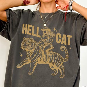 May include: Dark gray t-shirt with a gold graphic of a woman riding a tiger, with the words "HELL CAT" above the image. The shirt has a vintage, worn look.