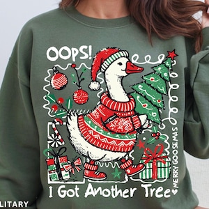 May include: A military green sweatshirt with a whimsical Christmas design. The graphic features a goose wearing a Santa hat and sweater, carrying a Christmas tree. Text includes "OOPS!" and "I Got Another Tree."