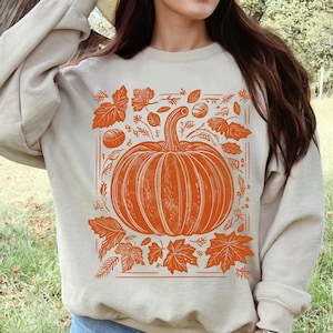 May include: Sand-coloured sweatshirt with an orange pumpkin design, leaves, and a rectangular border. The word "SAND" is printed at the bottom. Ideal for autumn and fall.