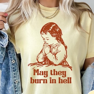 Puede incluir: Camiseta de color mantequilla con un gráfico marrón de un niño rezando y el texto "May they burn in hell". La camiseta tiene mangas cortas y un corte holgado.