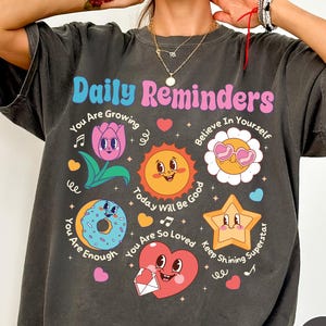 May include: Dark gray t-shirt with the text "Daily Reminders" in pink and blue. The shirt features colorful cartoon graphics of a flower, sun, donut, star, and heart with positive affirmations. The shirt is a casual, comfortable style.