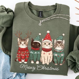 May include: Olive green sweatshirt with a graphic of four cats wearing holiday sweaters and hats. The cats are tan, black, and grey, with the text "Meowy Christmas" below. A silver necklace is draped on the sweatshirt.