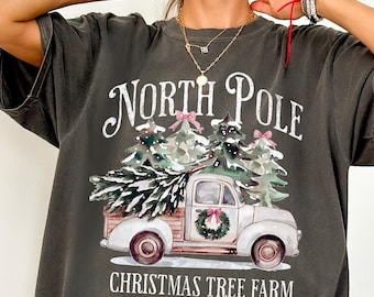 May include: Dark gray t-shirt featuring a vintage-style illustration. The design includes a white pickup truck carrying Christmas trees, with the text "NORTH POLE CHRISTMAS TREE FARM Always Fresh". The graphic has a watercolor effect.