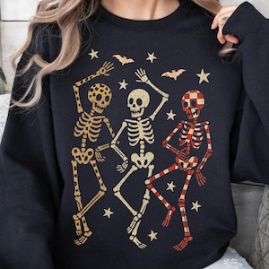 Puede incluir: Sudadera negra con tres esqueletos bailando en patrones dorados y a cuadros rojos, con estrellas y murci&eacute;lagos. La palabra "BLACK" est&aacute; impresa en la parte inferior. Un dise&ntilde;o festivo para Halloween.