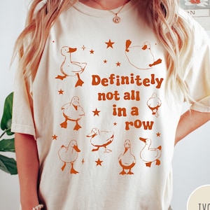May include: Ivory t-shirt with a graphic of ducks in a row and the text "Definitely not all in a row".