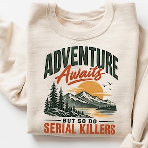May include: A cream-colored crewneck sweatshirt featuring a graphic print of a mountain landscape with pine trees, a sun, and birds. The text reads ADVENTURE Awaits BUT SO DO SERIAL KILLERS in dark green and orange lettering across the chest.
