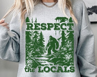 Puede incluir: Una sudadera gris con un estampado gráfico verde de un Bigfoot caminando por un bosque. El texto "Respect the Locals" está impreso debajo de la imagen.