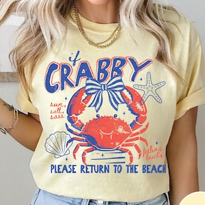 Pode incluir: Camiseta amarelo-claro com um desenho de caranguejo vermelho e azul e o texto "if Crabby, Please Return to the Beach". O design inclui uma concha e uma estrela do mar, com as palavras "sun, salt, sass" e "feeling beachy".