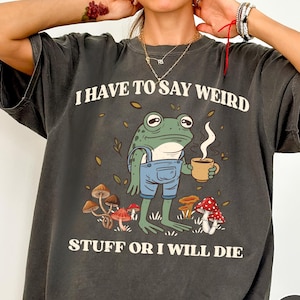 May include: Dark gray t-shirt with the text "I HAVE TO SAY WEIRD STUFF OR I WILL DIE." The shirt features a cartoon frog wearing blue overalls and holding a mug of coffee, surrounded by mushrooms and leaves.