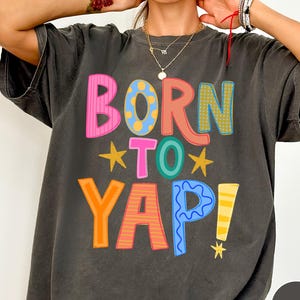 以下が含まれることがあります： ピンク、黄色、オレンジ、青などの鮮やかな色と様々なパターンで「BORN TO YAP!」というカラフルな文字がプリントされたチャコールグレーの半袖Tシャツ。小さな黄色い星と黄色い感嘆符がアクセントになっています。