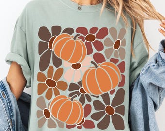 May include: A light green t-shirt featuring a fall-themed design. The design includes orange pumpkins and flowers in shades of brown, pink, and white. The shirt has short sleeves and a relaxed fit, perfect for autumn.