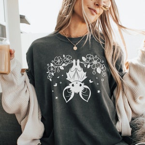 May include: A dark grey t-shirt featuring a white graphic of a bat hanging upside down with flowers and stars. The bat has a cute cartoon face.