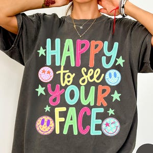 May include: A charcoal gray t-shirt featuring the colorful text HAPPY to see YOUR FACE in a mix of pink, blue, yellow, and orange. The design includes four smiley face icons with various patterns and small green stars scattered throughout the graphic.