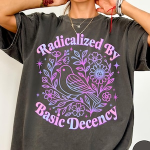 以下が含まれることがあります： 鳥と花のデザインが施されたダークグレーのTシャツ。「Radicalized By Basic Decency」の文字がピンクと青のグラデーションで描かれています。デザインはTシャツの前面中央に配置されています。