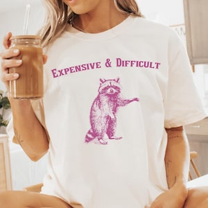 May include: A white t-shirt with the text "EXPENSIVE & DIFFICULT" printed in pink. A pink illustration of a raccoon is below the text. The t-shirt is labelled "IVORY".