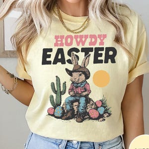 Puede incluir: Camiseta amarilla clara con las palabras "HOWDY EASTER" en rosa y negro. El gr&aacute;fico presenta un conejo con sombrero de vaquero, sentado en una roca con huevos de Pascua y un cactus. Un c&iacute;rculo amarillo representa el sol.