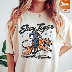 May include: Ivory-colored t-shirt featuring a graphic of a cowboy riding a tiger, with the text "Easy Tiger" above. The cowboy is in blue, the tiger is orange and black, and blue stars are scattered around the text. The shirt is a relaxed fit.